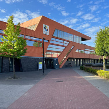 Externe foto van een modern kantoorgebouw met oranje lambrisering en donkere baksteen. De ingang is gemarkeerd met het nummer 7 en een logo met de tekst "Vitalogie PLUS" is zichtbaar. De stoep leidt naar de ingang, met kleine bomen aan weerszijden, onder een blauwe lucht met wolken.