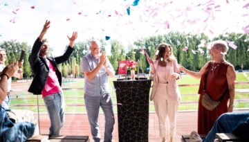 Een feestelijke scène buiten op een terras met uitzicht op een atletiekbaan, waarbij roze en blauwe confetti neerdaalt. Een lachende vrouw in een lichtroze pak staat centraal naast een zwarte statafel met daarop een rood boek, wat duidt op een boeklancering. Links van haar juicht een man in een roze shirt met zijn armen in de lucht, terwijl andere omstanders blij toekijken en klappen.