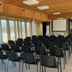 Een lege presentatieruimte met een houten plafond en grote ramen aan de zijkant. Zwarte stoelen staan in rijen opgesteld, gericht op een projectiescherm. Rechtsachter staat een flip-over en een dartbord-achtig spel.