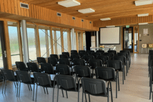 IMG_8839 Een lege presentatieruimte met een houten plafond en grote ramen aan de zijkant. Zwarte stoelen staan in rijen opgesteld, gericht op een projectiescherm. Rechtsachter staat een flip-over en een dartbord-achtig spel.