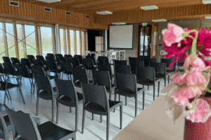 IMG_8843 Een lege presentatieruimte met houten plafond en grote ramen. Zwarte stoelen staan in rijen gericht op een projectiescherm. Op de voorgrond staat een vaas met roze en rode bloemen.