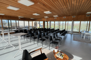 IMG_8844 Een lichte presentatieruimte met een houten plafond en grote ramen. Zwarte stoelen staan in rijen opgesteld voor een podium. Rechts en links op de voorgrond zijn tafels en een zitgedeelte zichtbaar.