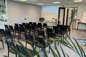 IMG_8851 Een lege presentatieruimte met rijen zwarte stoelen, een grijze vloer en een witte muur. Achterin is een projectie zichtbaar, een deuropening en een kleine kitchenette.