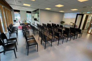 IMG_8853 Een lege presentatieruimte met rijen zwarte stoelen, een grijze vloer en een witte muur. Achterin is een projectie zichtbaar, een deuropening en een kleine kitchenette.