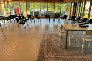 IMG_9693 Een lichte, moderne vergaderruimte met houten lambrisering en grote ramen. Zwarte stoelen staan in een cirkel op de glanzende vloer. Rechts staat een witte tafel op een vloerkleed, en op de achtergrond zijn posters van 'Vitalogie' zichtbaar.