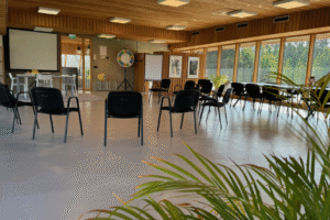 IMG_9697 Een lichte vergaderruimte met houten lambrisering en grote ramen die uitkijken op groen. Zwarte stoelen staan in een halve cirkel, en in de hoek staat een projectiescherm en een flip-over.