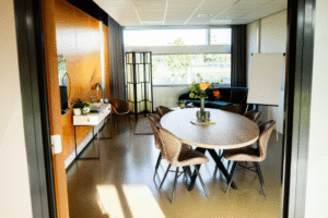 Vitalogisch -17 Een lichte, moderne vergaderruimte gezien door een deuropening, met een ovale houten tafel, bruine stoelen, een flip-over en een zithoek met uitzicht op buiten.