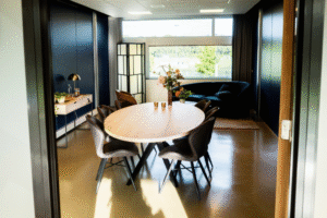 Vitalogisch -19 Uitzicht door een deuropening op een luxe vergader- of loungeruimte met een ovale houten tafel, fluwelen stoelen en een donkerblauwe bank, met uitzicht op een groen landschap.