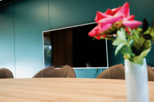 Vitalogisch -23 Close-up van een houten vergadertafel met een vaas kunstbloemen en op de achtergrond een groot beeldscherm tegen een turquoise wand.