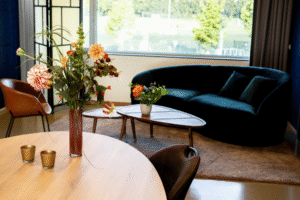 Vitalogisch -28 Een stijlvolle zithoek in een kantoorruimte met een donkergroene bank en een salontafel, gescheiden van een ronde vergadertafel door een bloemstuk.