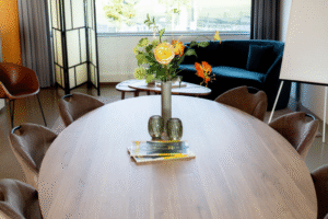 Vitalogisch -29 Ovale houten vergadertafel met bloemstuk en tijdschriften, omringd door stoelen. Op de achtergrond is een zithoek en een flip-over zichtbaar.