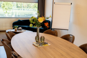 Vitalogisch -30 Interieur van een lichte vergaderkamer of lounge met een ovale houten tafel, bruine stoelen en een donkerblauwe bank, met uitzicht op een sportveld.